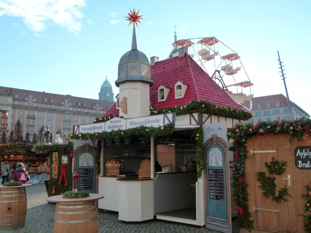 Kleines rotes Dachgebäude mit einem Riesenrad davor, umgeben von Festschmuck, Menschen und Gegenständen, vor Gebäuden, Bäumen und einem bewölkten Himmel während des Oktoberfests in München, Deutschland.