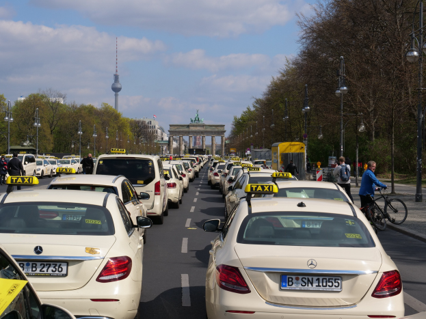 Eine lange Reihe von Taxis geparkt entlang einer belebten Straße in Berlin, Deutschland, mit Fahrradfahrern und Fußgängern auf dem Gehweg, flankiert von Bäumen und Laternen, und Gebäuden, einem Bogen und einem Turm im Hintergrund unter einem bewölkten Himmel.