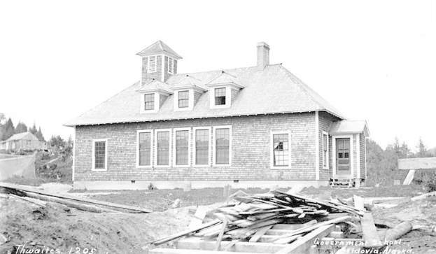 Schwarzes und weißes Foto eines zweistöckigen Gebäudes mit Fenstern und Türen, umgeben von Bäumen unter einem klaren Himmel, beschriftet mit "Regierungs-Schule, Thwaites, 1905" mit Holzplanken unten.