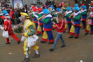 Eine Gruppe von Menschen in bunten Kostümen marschiert durch eine Straße, spielt Musikinstrumente und hält Blumensträuße, mit Zuschauern und Gebäuden im Hintergrund.