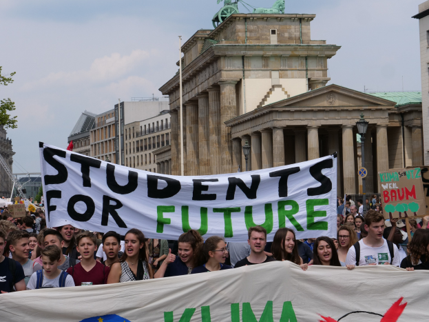 Eine Gruppe von Schülern marschiert in Berlin, hält ein buntes "Students for Future"-Schild gegen eine Kulisse aus Gebäuden, Bäumen und Himmel.