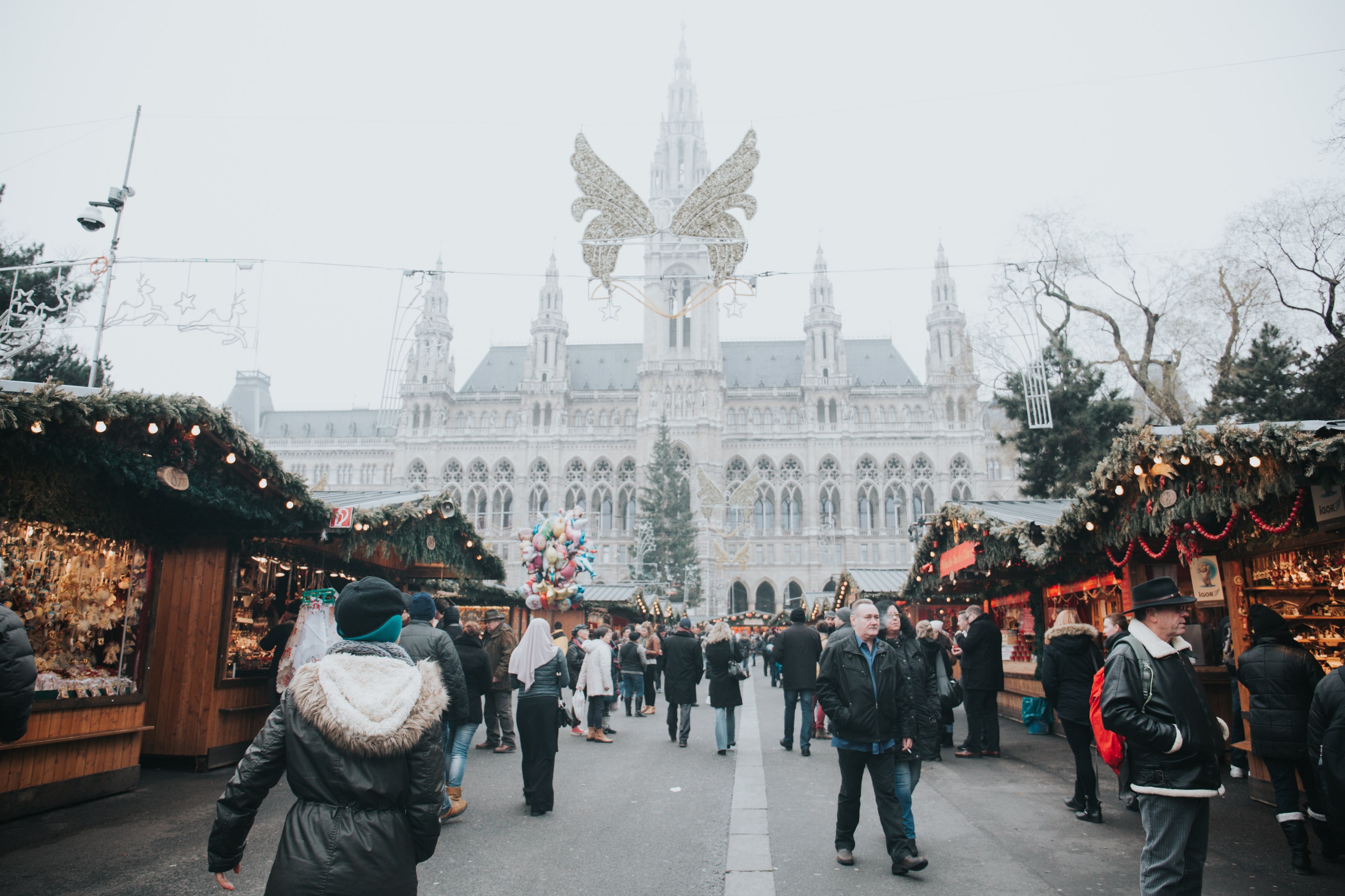 Ein lebendiger Weihnachtsmarkt in Wien, Österreich, mit Menschen, die herumlaufen, Ständen mit Lichtern und festlichen Dekorationen, einem Gebäude mit Fenstern im Hintergrund, Bäumen und einem klaren blauen Himmel.