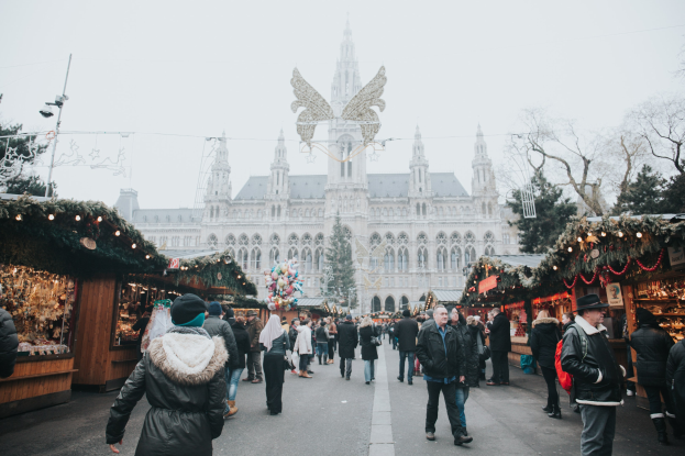 Ein lebendiger Weihnachtsmarkt in Wien, Österreich, mit Menschen, die herumlaufen, Ständen mit Lichtern und festlichen Dekorationen, einem Gebäude mit Fenstern im Hintergrund, Bäumen und einem klaren blauen Himmel.