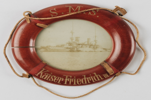 Rotes Rettungsfloß mit 'USS Kaiser Friedrich III'-Text und Battleship-Bild, an einer weißen Oberfläche mit Seilen befestigt.
