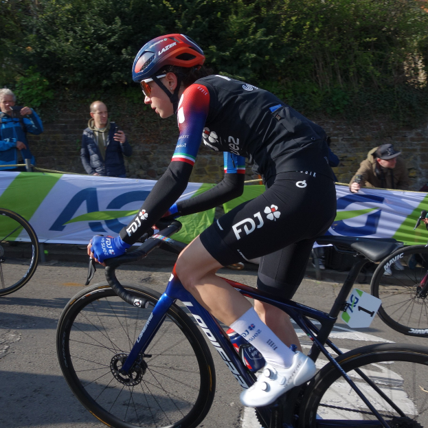 Eine professionelle Radsportlerin, Katie Archibald, fährt mit Helm, Brille, Handschuhen und Schuhen ein Fahrrad auf einer Straße neben einer Menge, mit einem Banner, einer Wand, Bäumen und einem Gebäude im Hintergrund.