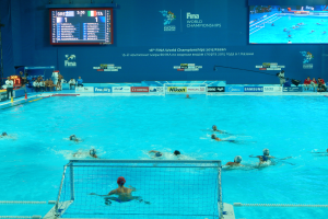 Eine Gruppe von Menschen, die Wasserball in einem großen Pool mit einem Netz spielen, Zuschauer sitzen links und ein Bildschirm im Hintergrund zeigt Texte und Logos für die FINA-Weltmeisterschaften 2016 in Kazan.