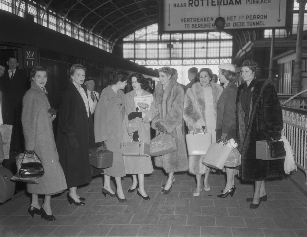 Schwarzes und weißes Bild einer Gruppe von Frauen, die an einem Bahnhof stehen, jede hält eine Tasche, mit einem Zug links und Geländern und einer Tafel rechts.