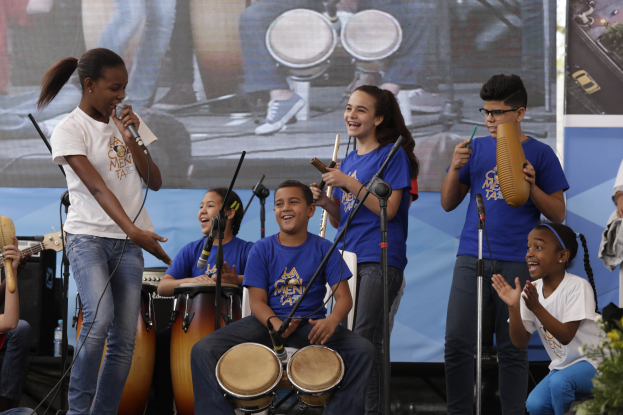 Kinder, die auf der Bühne mit Musikinstrumenten spielen, eine Frau mit einem Mikrofon, Mikrofonständer, eine Banner im Hintergrund und Topfpflanzen auf der rechten Seite.