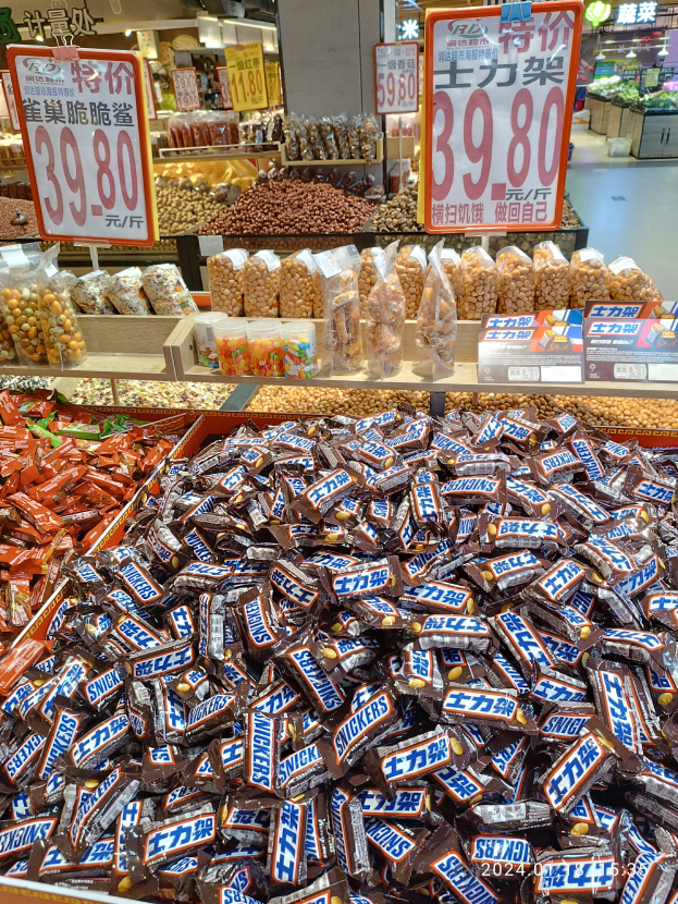 Eine Vitrine in einem Geschäft, gefüllt mit verschiedenen Sorten von Süßigkeiten, darunter Snickers, mit Lebensmitteln auf Regalen, Text- und Zahlentafeln, Deckenleuchten und anderen Gegenständen im Hintergrund.