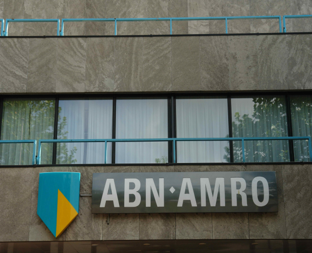 Ein modernes Bürogebäude mit "ABN AMRO"-Schriftzug, einer Tafel, die seinen Status als Hauptsitz anzeigt, Geländern und Vorhangfenstern, die auf eine EU-Strafe von 1,5 Milliarden Dollar hinweisen.