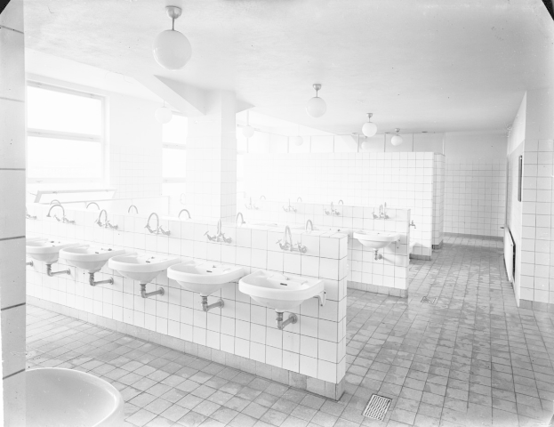 Schwarz-weiß-Foto einer öffentlichen Toilette mit Waschbecken, Wasserhähnen, Wänden, Beleuchtung und Fenstern.