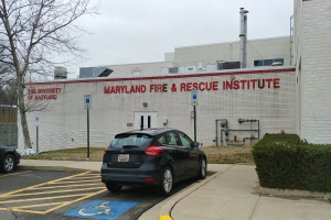 Ein Gebäude des Maryland Fire & Rescue Institute mit Text darauf, ein Auto davor geparkt, Schilder, Pfosten, Rohre und andere Gegenstände im Vordergrund, Pflanzen rechts, ein Baum links und der Himmel oben sichtbar.
