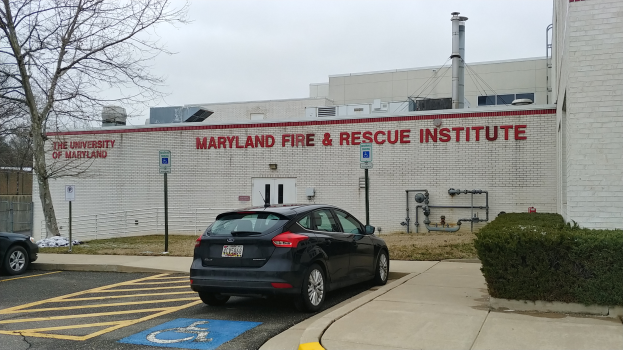 Ein Gebäude des Maryland Fire & Rescue Institute mit Text darauf, ein Auto davor geparkt, Schilder, Pfosten, Rohre und andere Gegenstände im Vordergrund, Pflanzen rechts, ein Baum links und der Himmel oben sichtbar.