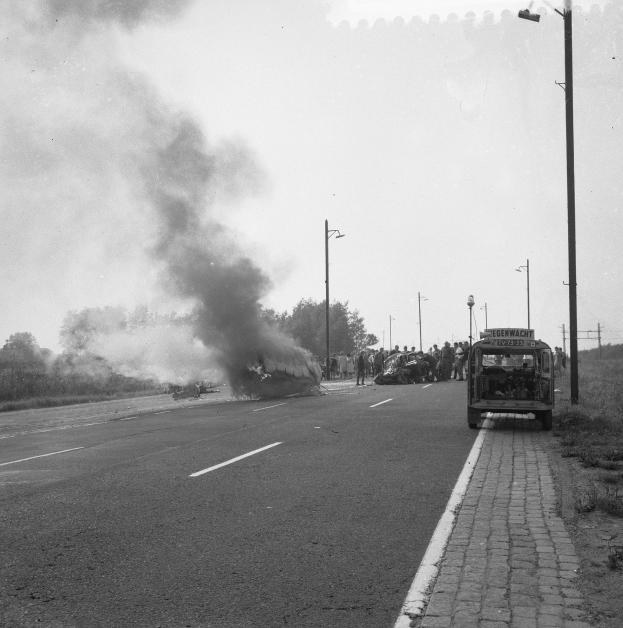 Schwarz-weiß-Foto eines brennenden Busses auf einer Straße mit ein paar Menschen in der Nähe und Strommasten, Bäumen und Himmel im Hintergrund.