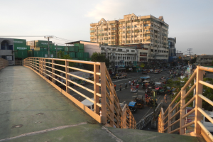 Eine Fußgängerbrücke mit Geländern überspannt eine vielbefahrene Straße mit vielen Fahrzeugen und Fußgängern, im Hintergrund sind Gebäude, Bäume, Strommasten und ein bewölkter Himmel zu sehen.