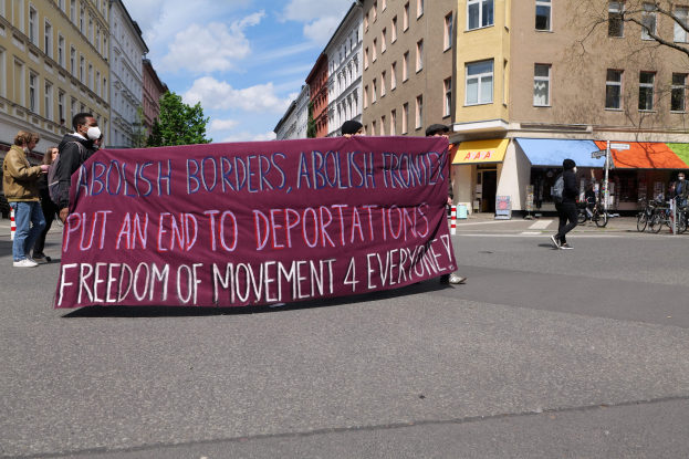 Eine Gruppe von Menschen marschiert auf einer Straße, hält ein Banner mit der Aufschrift "Abolish Borders, Abolish Frontiers, Put an End to Deportations, Freedom of Movement 4 Everyone", mit Gebäuden, Bäumen, Fahrrädern und einem bewölkten Himmel im Hintergrund.