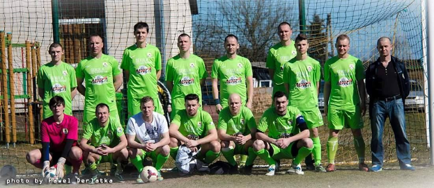 Eine Gruppe von Männern in grünen T-Shirts steht auf einem Fußballfeld mit einem Ball davor, einige in der Hocke, in der Nähe eines Torpfostens mit Bäumen, Gebäuden und Fahrzeugen im Hintergrund.