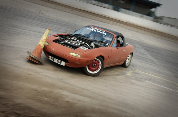 Eine Person fährt ein rotes Auto auf einer Rennstrecke und überholt einen Verkehrskegel, während der Hintergrund unscharf ist, was auf hohe Geschwindigkeit hinweist.