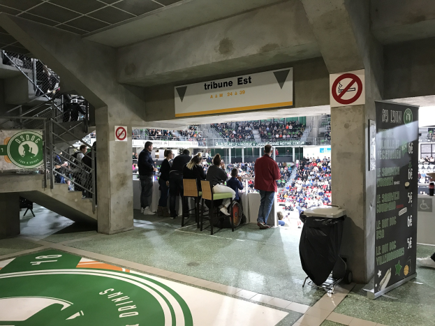 Gruppe von Menschen vor einer sitzenden Menge in einem Stadion mit Schildern, die "Boston Celtics vs New York Jets" lesen, ein Müllcontainer, eine Tafel mit Text, eine Treppe mit Geländern und eine Decke mit Deckenleuchten.
