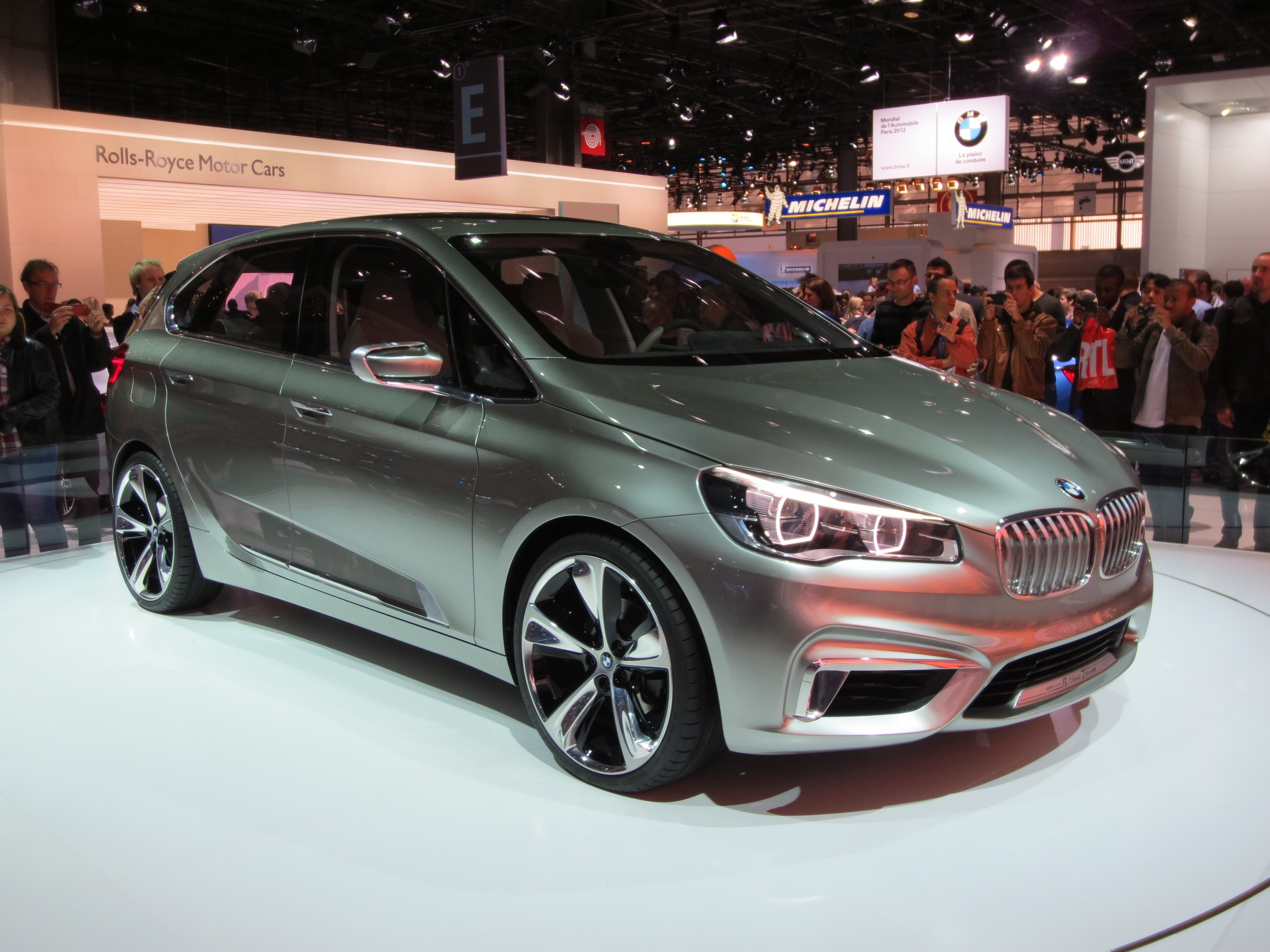 BMW 2er Active Tourer Concept auf der IAA in Frankfurt gezeigt, umgeben von Menschen, die es fotografieren, unter Deckenlampen mit Texttafeln im Hintergrund.