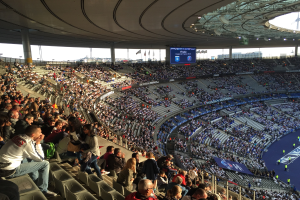 Großes Publikum sitzt im Allianz Arena Stadion und schaut ein Fußballspiel mit einer Bühne, Fahnen und einem Bildschirm im Hintergrund.