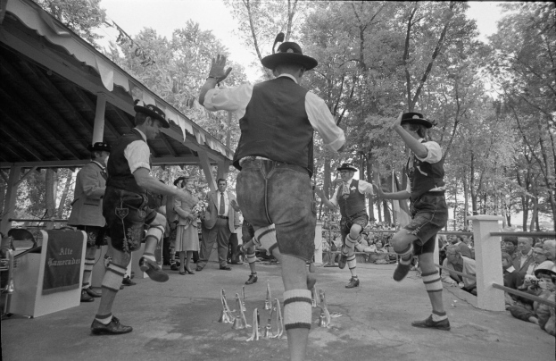 Eine Gruppe von Männern in traditioneller bayrischer Tracht, die auf einer Bühne vor einem gemischten Publikum tanzen, vor einem Hintergrund aus Bäumen und einer Hütte dargestellt in Schwarz-Weiß.