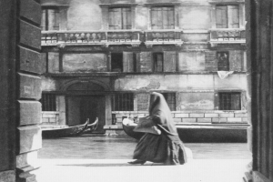 Eine Frau spaziert auf einer Straße in Venedig, Italien, mit einem Gebäude im Hintergrund und zwei Booten auf dem Wasser, dargestellt in Schwarz-Weiß.