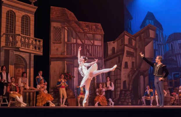 Zwei Tänzer performen auf der Bühne im Royal Opera House in London, mit einem Publikum im Vordergrund, das auf Stühlen sitzt und steht, und einer Kulisse aus Gebäudezeichnungen.