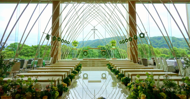 Eine Hochzeitszeremonie in einem großen Innenraum mit halbrunder Sitzordnung, Blumenschmuck und einem Hintergrund mit Bäumen, Hügeln und einem klaren Himmel.