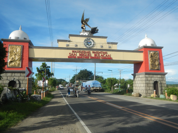 Eingang einer Stadt mit einem Tor mit Text, Skulpturen und einer Statue oben drauf, flankiert von Bäumen, Fahrzeugen, Schildern, Strommasten mit Drähten und einem bewölkten Himmel.