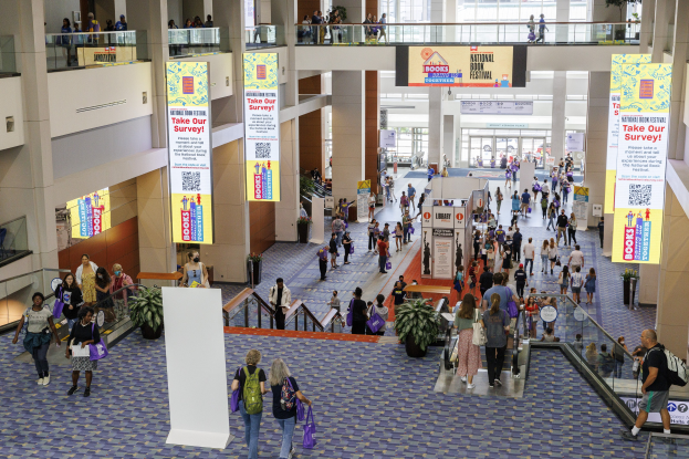 Eine große Menschenmenge bewegt sich durch ein großes Gebäude mit Bannern, Rolltreppen, Geländern, Säulen, Texttafeln, Glastüren und Topfpflanzen, einige tragen Taschen, wahrscheinlich nehmen sie am Nationalen Buchfestival teil.