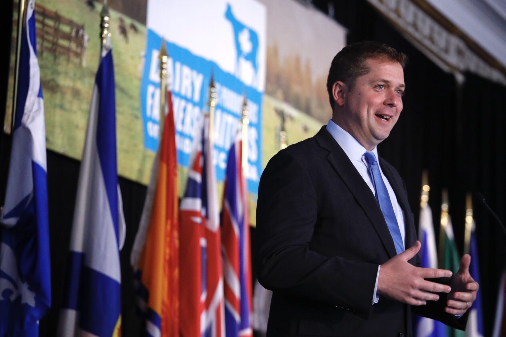 Ein Mann in Anzug und Krawatte spricht an einem Podium mit Mikrofonen, umgeben von Flaggen und einem Bildschirm, wahrscheinlich spricht er über eine Regierungszahlung von 1,5 Milliarden Dollar für die Milchindustrie.