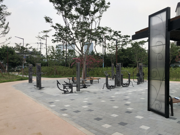 Outdoor park with fitness equipment, benches, trees, grass, poles, lights, wires, buildings, and sky.