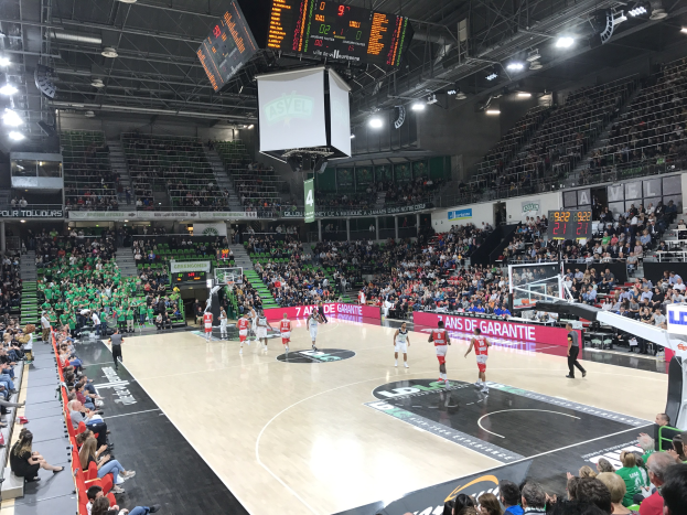 Ein Basketballspiel in einer großen Arena mit Zuschauern auf den Tribünen, Anzeigetafeln mit 'Asvel vs Galatasaray' und einer Decke mit Lampen und einem Bildschirm.