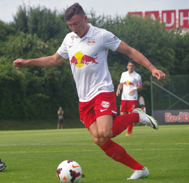 Professioneller Fussballspieler, der einen Ball auf einem Rasenfeld kickt, mit "RB Leipzig" im Hintergrund und einem klaren blauen Himmel.