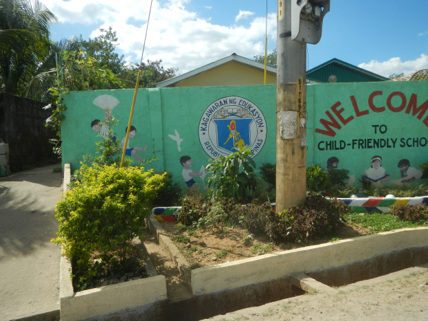 Eine grüne Wand mit einem Schild, auf dem "Willkommen in der kinderfreundlichen Schule" steht, umgeben von Häusern, Bäumen, Pflanzen und einem Mast mit einer Lampe, unter einem bewölktem Himmel.