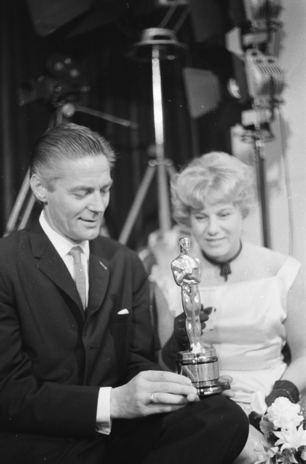 Ein Mann in einem schwarzen Blazer, weißem Hemd und Krawatte hölt eine Oscar-Statue, während er neben einer Frau in Abendgarderobe sitzt, die einen Blumenstrauß hölt, mit weicher Beleuchtung im Hintergrund.