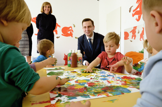Gruppe von Kindern bei einer Malaktivität an einem Tisch mit farbigen Händen, Papieren und Flaschen, beaufsichtigt von einer lächelnden Frau, mit Kunstwerken an der Wand im Hintergrund.
