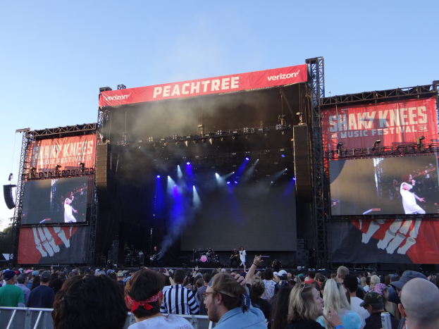 Eine große Menschenmenge vor einer hell erleuchteten Bühne beim Shaky Knees Music Festival, auf der Musiker hinter Bannern spielen, mit einem Geländer im Vordergrund und einem klaren blauen Himmel im Hintergrund.