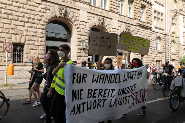 Eine Gruppe von Menschen geht eine Straße entlang, einige halten Schilder und Banner, andere fahren Fahrräder, mit einem Gebäude im Hintergrund, das Fenster, Bögen, Säulen und Skulpturen aufweist, Bäume und ein Laternenmast, an der Protestaktion gegen die Anti-Kriegs-Bewegung in Berlin teilnehmen.