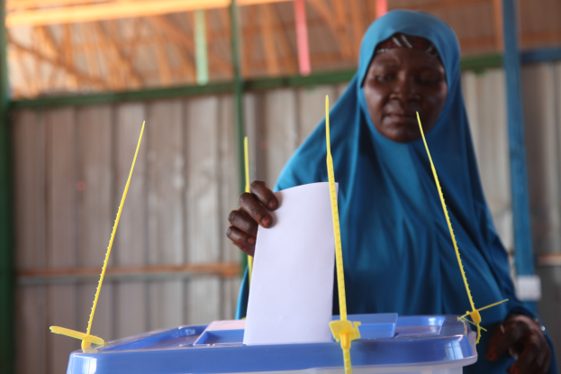 Frau in blauem Kopftuch hält ein Blatt Papier in der Hand und steht vor einer Kiste mit herausragenden Stöcken, vor einer Wand und einem Schuppen, was auf eine Wahllokalität hinweist.