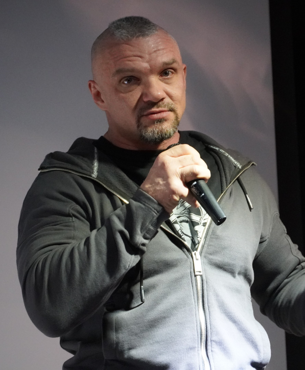 Ein Mann in einer schwarzen Kapuzenjacke, schwarzem Anzug und weißem Hemd, der selbstbewusst vor einer Wand steht und ein Mikrofon hält.