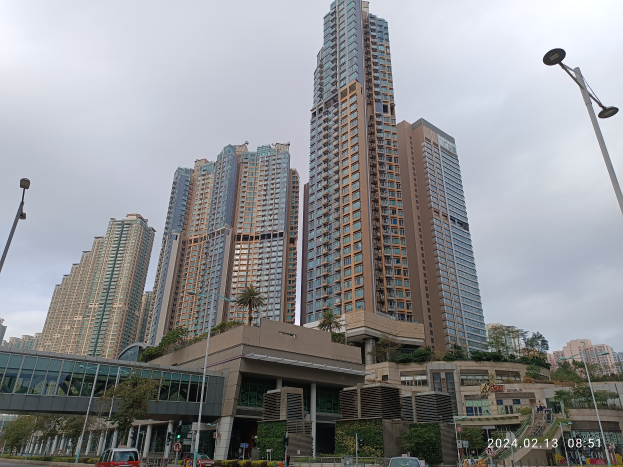 Eine Stadtstraße mit hohen Gebäuden, Fahrzeugen, Laternen, einer Brücke, Bäumen und einem bewölkten Himmel, mit einem prominenten Wohngebäude im Vordergrund.