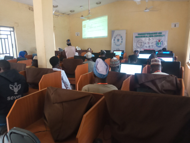 Gruppe von Menschen, die an Computern in einem Seminarräumen mit Deckenventilatoren, einem Bildschirm, einer Tafel und Fenstern auf der linken und rechten Seite sitzen.