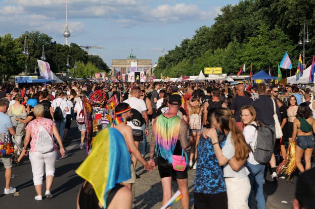 Eine große Menge von Menschen mit Taschen und Fahnen marschiert auf einer Straße in Berlin, Deutschland, vorbei an Zelten, Bäumen, Laternenmästen, einem Tor und einem Turm.