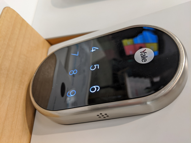 Nahaufnahme eines Yale Smart Lock-Geräts auf einem weißen Tisch mit einem hölzernen Objekt links daneben.