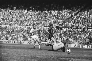 Schwarzes und weißes Foto von Männern, die auf einem Feld Fußball spielen, mit Zuschauern im Hintergrund und einer "Bundesliga 1978-79 Ajax 1-1 Hannover 96"-Fahne.