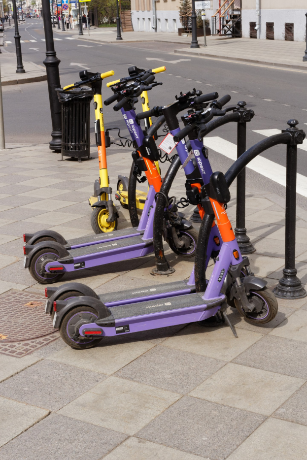 Eine Reihe von E-Scootern, die entlang einer Straße geparkt sind, mit einem Müllcontainer im Vordergrund und Gebäuden, Bäumen und Fahrzeugen im Hintergrund.