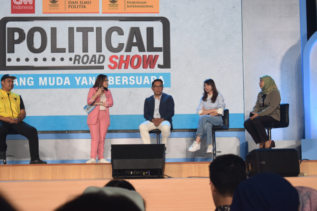 Eine Gruppe von Menschen sitzt auf einer Bühne mit einer Frau, die ein Mikrofon in der Mitte hält, einer Zuschauermenge im Vordergrund und einem Banner mit der Aufschrift 'Politischer Roadshow 2019' im Hintergrund.