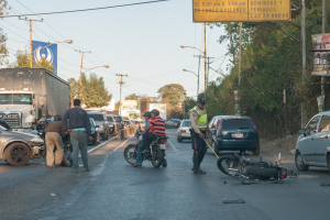 Eine Gruppe von Menschen steht um ein verunglücktes Motorrad auf der Straßenseite mit mehreren Fahrzeugen, darunter ein Lastwagen, und einer Hintergrund von Bäumen, Masten, Lichtern und Schildern unter einem Himmel.